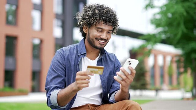 Happy young man enters a credit card data on a mobile phone while sitting on a bench on the street near a modern building. Smiling handsome male shopping online, enjoy e-commerce e-shopping. Close up