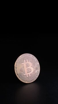 Vertical video of a golden bitcoin cryptocurrency coin standing on a reflective dark surface in a studio setting while moving lights create glowing reflections and shimmering metallic highlights, symb