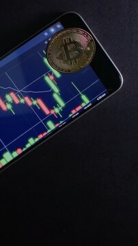 Vertical video of a smartphone positioned at an angle against a dark background while displaying a bitcoin trading chart with a golden cryptocurrency coin placed on the screen, featuring negative spac