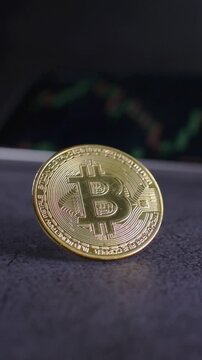 Vertical close up video of a golden bitcoin cryptocurrency coin standing on a granite surface while a blurred smartphone in the background shows a financial trading chart, symbolizing blockchain techn