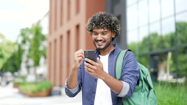 Satisfied happy male student received great news on mobile phone standing on street in campus space near university building. Smiling joyful handsome man reads a positive good message on smartphone
