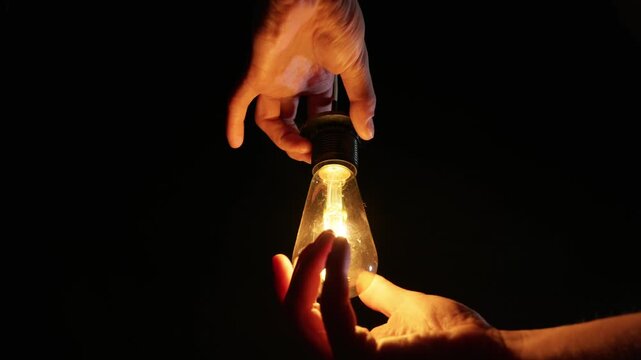 Two hands gently screw a flickering vintage light bulb into a hanging electrical socket. The warm glowing internal filament provides the only illumination in the pitch black environment.