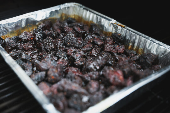 Pork Belly Burnt Ends cooked until black on a pellet grill smoker. The delicious morsels of fatty pig meat are resting in a tinfoil pan ready to eat for dinner. 