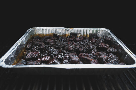 Pork Belly Burnt Ends cooked until black on a pellet grill smoker. The delicious morsels of fatty pig meat are resting in a tinfoil pan ready to eat for dinner. 