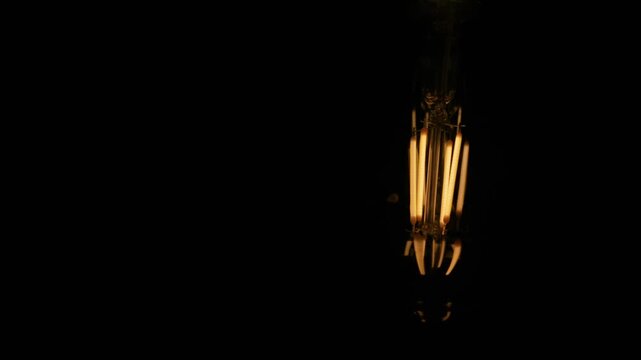 A vintage LED filament light bulb glows brightly against a pitch black background. Intricate wire structures radiate a warm amber light inside the dusty glass casing. It shines in isolation.