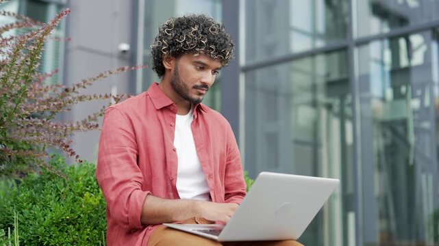 Confident young businessman uses a laptop while sitting on a bench near business office building. Handsome man working, chatting online with a client, typing an email message or browsing web. Close up