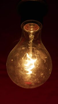 An old incandescent light bulb glows brightly against a dark background. The warm light illuminates the delicate filament and dust on the glass. The atmosphere is nostalgic and moody.