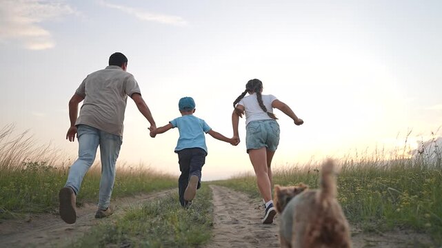 Family runs holding hands on path with dog following at sunset. Father with children running together outdoors. Family and dog enjoy evening run. Children hold hands while running with father and pet.