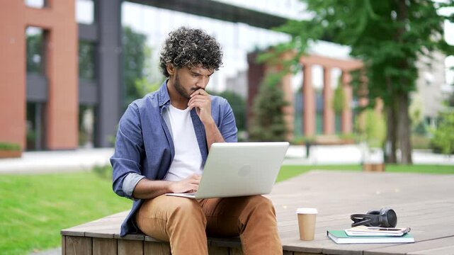Thoughtful male student using laptop while sitting on a bench in campus space near the university building. Freelancer thinks about solving the problem while working on the computer, found the answer