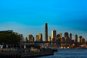 Fototapeta premium The massive skyscrapers of Brooklyn rise prominently behind the steel structure of the Williamsburg Bridge as warm golden light hits the urban horizon during sunset.