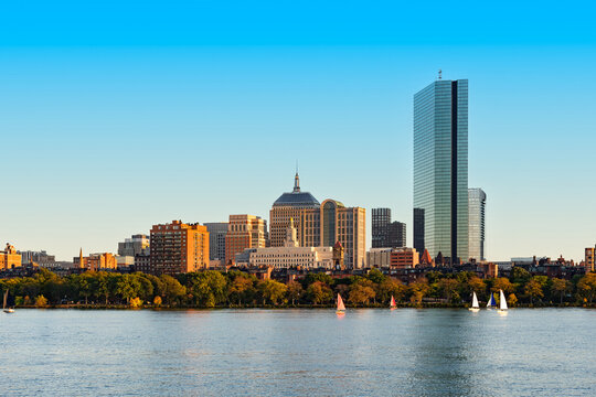 Multiple small sailboats navigate the calm waters of the Charles River, which flows in front of the diverse architectural skyline of Boston, Massachusetts.