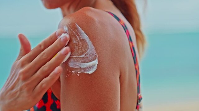 Young woman applying moisturizing sunscreen lotion on a shoulder with a beautiful ocean in the background. Skin care sun protection. Closeup