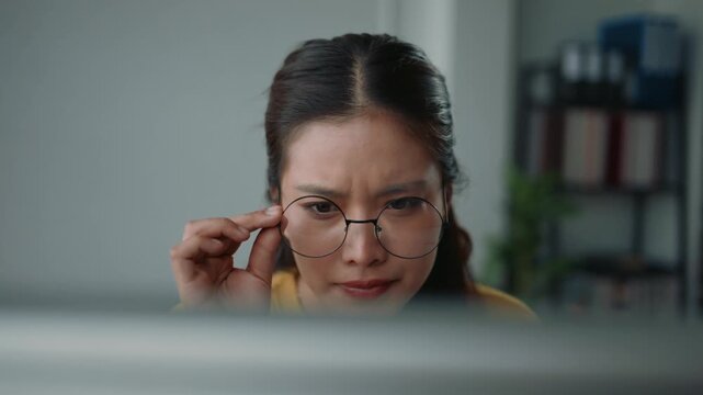 Young asian woman with glasses experiencing blurry vision and eye strain while working on her computer in the office, feeling tired