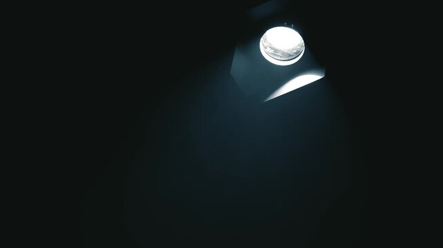 High-contrast static shot of a cold white spotlight fixture on a dark ceiling. Subtle stage smoke and haze slowly drift through the crisp light beam. Perfect minimalist background