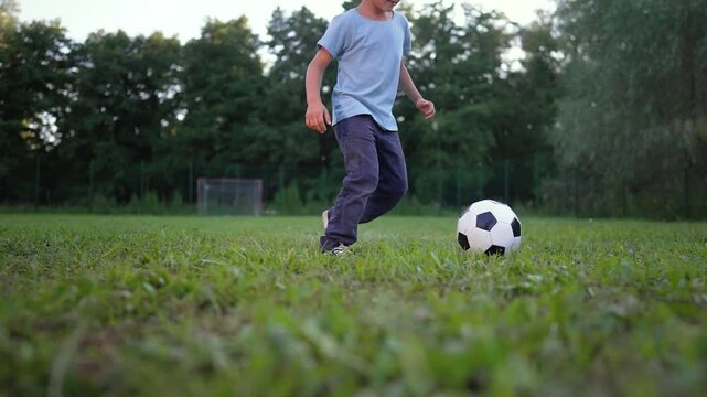 Boy runs with soccer ball on grass field at park during evening. Child plays football outdoors near goal. Kid dribbles soccer on grass. Boy with ball on field. Child running with football at park.