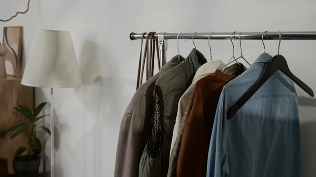 No people shot of several shirts and jackets hanging on metal clothing stand next to floor lamp and potted plant, with white wall in background