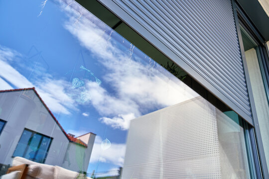 Grey exterior roller shutter is partially open above a large glass window on a modern house facade. Home security, privacy, sun protection and residential exterior