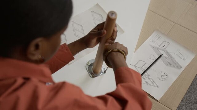 Over the shoulder view of African American young woman screwing table leg to wooden top while assembling furniture with help of paper instructions