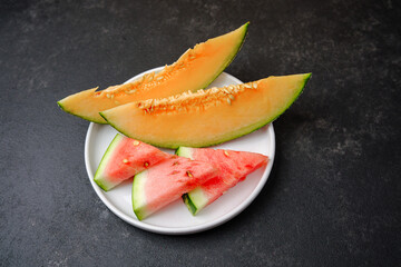 Slices of ripe cantaloupe and watermelon on a white plate against a dark textured background.