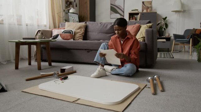Black young woman pulling up sleeve of her shirt, reading table assembly instructions on paper and examining wooden leg while sitting on floor in living room