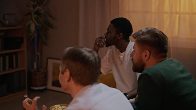 Medium shot of biracial friends sitting in living room and watching football, rooting for team