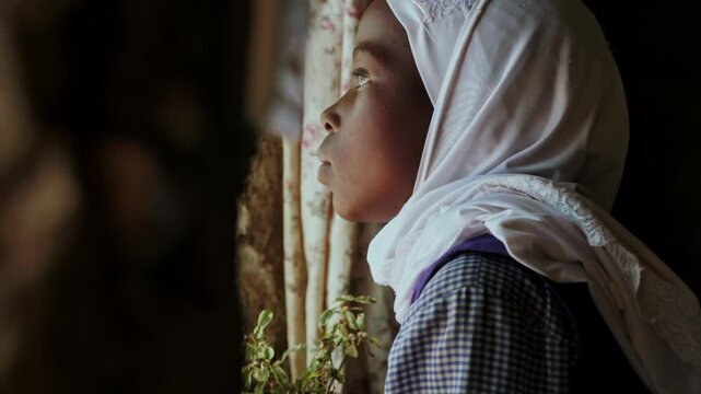 A young girl in a white headscarf looks outside through an old barred window, reflecting childhood, education, rural home life, quiet hope, innocence and thoughtful curiosity indoors.