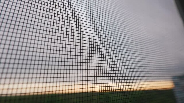A detailed close-up focuses on the fine texture of a mosquito screen on a window frame overlooking a blurred sunset landscape. Concept of home protection, insect prevention, and pest control.