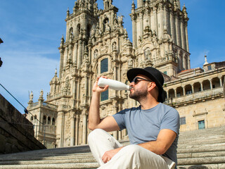 Fototapeta premium Man in a fedora hat sitting on stairs drinking from a reusable water bottle at Santiago Cathedral.