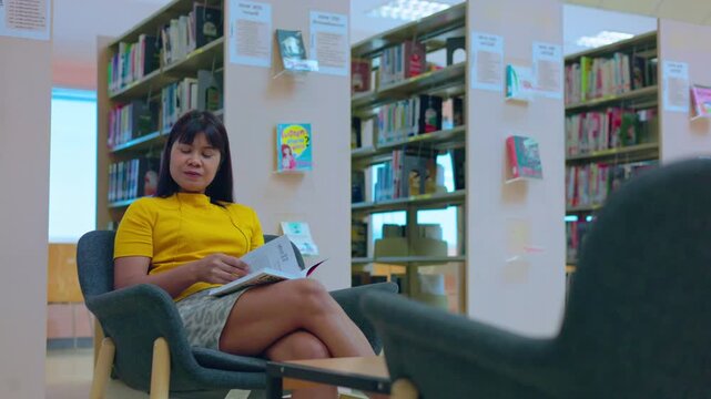 Asian woman sitting on a comfortable chair and reading a book in a modern library. Education, learning, and self-improvement concept with bookshelves background.