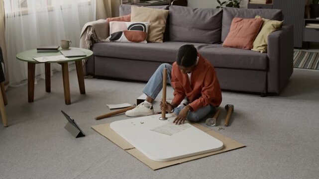 African American young woman assembling coffee table with help of paper instructions while sitting on carpeted floor of modern living room