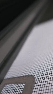 Macro detail reveals the geometric grid of a white mosquito net mounted against a dark blurred window frame. Concept of insect protection, home safety, and abstract textural backgrounds.