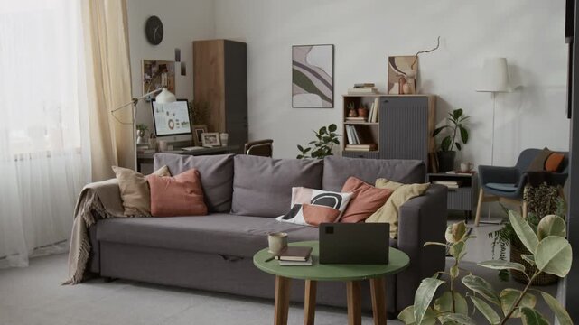 Interior of living room featuring large grey couch with throw pillows, green coffee table with laptop and books, as well as computer desk, cabinets and armchair in background