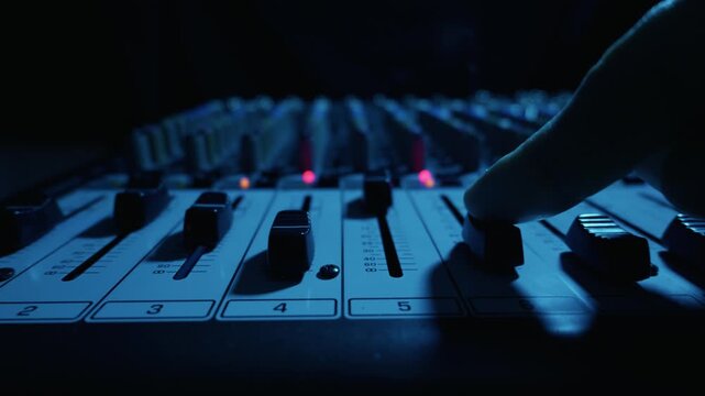 Hand adjusting a channel fader on a professional audio mixing console illuminated by green light in a dark studio. This illustrates the concept of professional sound engineering.