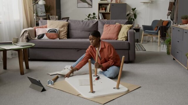 Black young woman attaching wooden legs to white tabletop using screwdriver while sitting on floor of modern living room next to digital tablet
