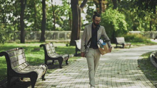 Fired Businessman Walking in City Park Carrying Box with Personal Office Belongings. Career Failure and Professional Crisis