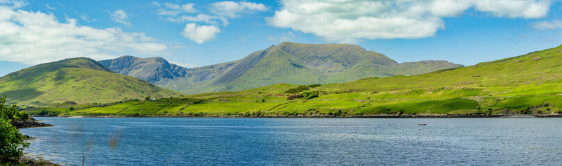 Fototapeta premium Green mountain slopes rise above Killary Fjord under bright skies in western Ireland. This image is a scenic landscape focused more on the fjord lush hillsides than the water.