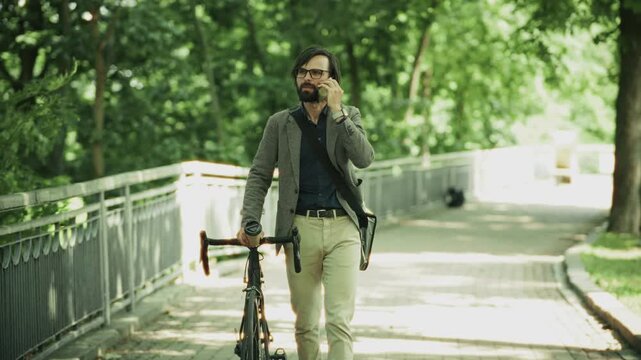 Businessman Walking with Bicycle and Talking on Smartphone in City Park. Eco Friendly Transportation