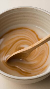 A close-up macro shot of mixed coffee and milk in a speckled ceramic bowl, with a wooden spoon resting in the swirling creamy beverage.