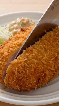 A close-up macro shot of a freshly fried breaded pork cutlet being sliced with a knife, served with coleslaw and shredded cabbage on a white ceramic plate.