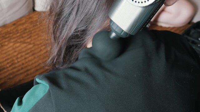 Woman applies a percussion massage gun to her shoulder muscles to relieve tension while sitting indoors. Concept of home wellness, muscle recovery, percussive therapy, and self care for health