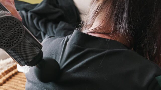 A woman is receiving a massage on her upper back and shoulder using a handheld percussion massage gun. The device provides deep tissue relief for muscle tension in a cozy domestic environment