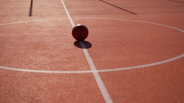 Basketball lies in center of basketball court on marking line on sunny day. Sports equipment before start of game. Worn ball on orange rubberized surface on arc pull in shot.