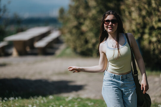 Young woman wearing sunglasses and a tank top walks through a sunny park path. She strolls casually carrying a shoulder bag and smiling slightly.