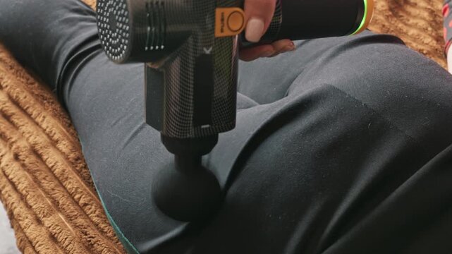 A close-up view shows a woman using a percussive massage gun on her thigh while lying down. The device vibrates against black leggings to relieve muscle tension and soreness after exercise.