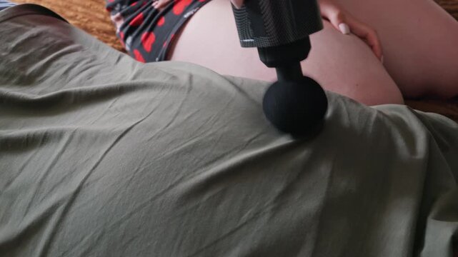 A person uses a black percussion massage gun on their partner back covered by an olive green shirt. The modern handheld device vibrates rapidly to relieve muscle tension at home.