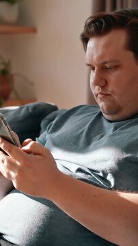 Vertical video: Reclining man in teal tee and black sweats tapping smartphone on sofa near plants