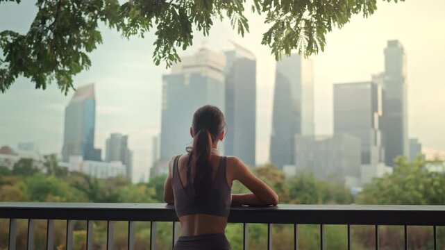 Back view of healthy Asian woman in sportswear standing at park balcony looking at modern city skyscrapers morning jog wellness routine and urban fitness lifestyle concept