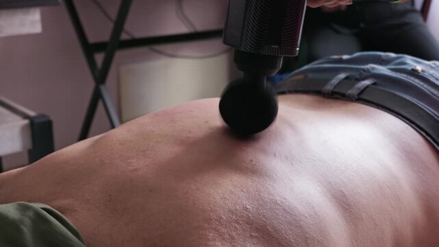 Man lies face down while receiving a deep back massage with a black percussion massage gun. Concept of muscle recovery and home physical therapy.