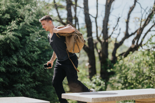 A young man runs carrying a backpack and smartphone in a leafy outdoor area. He moves briskly past stone benches under sunlit trees.