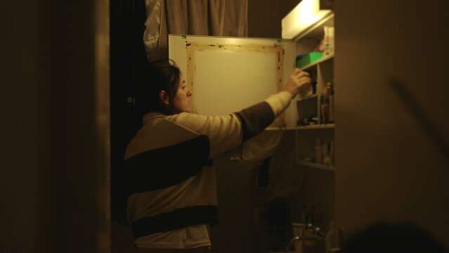 Woman standing in bathroom opening medicine cabinet and reaching for personal care products, everyday hygiene routine in home interior